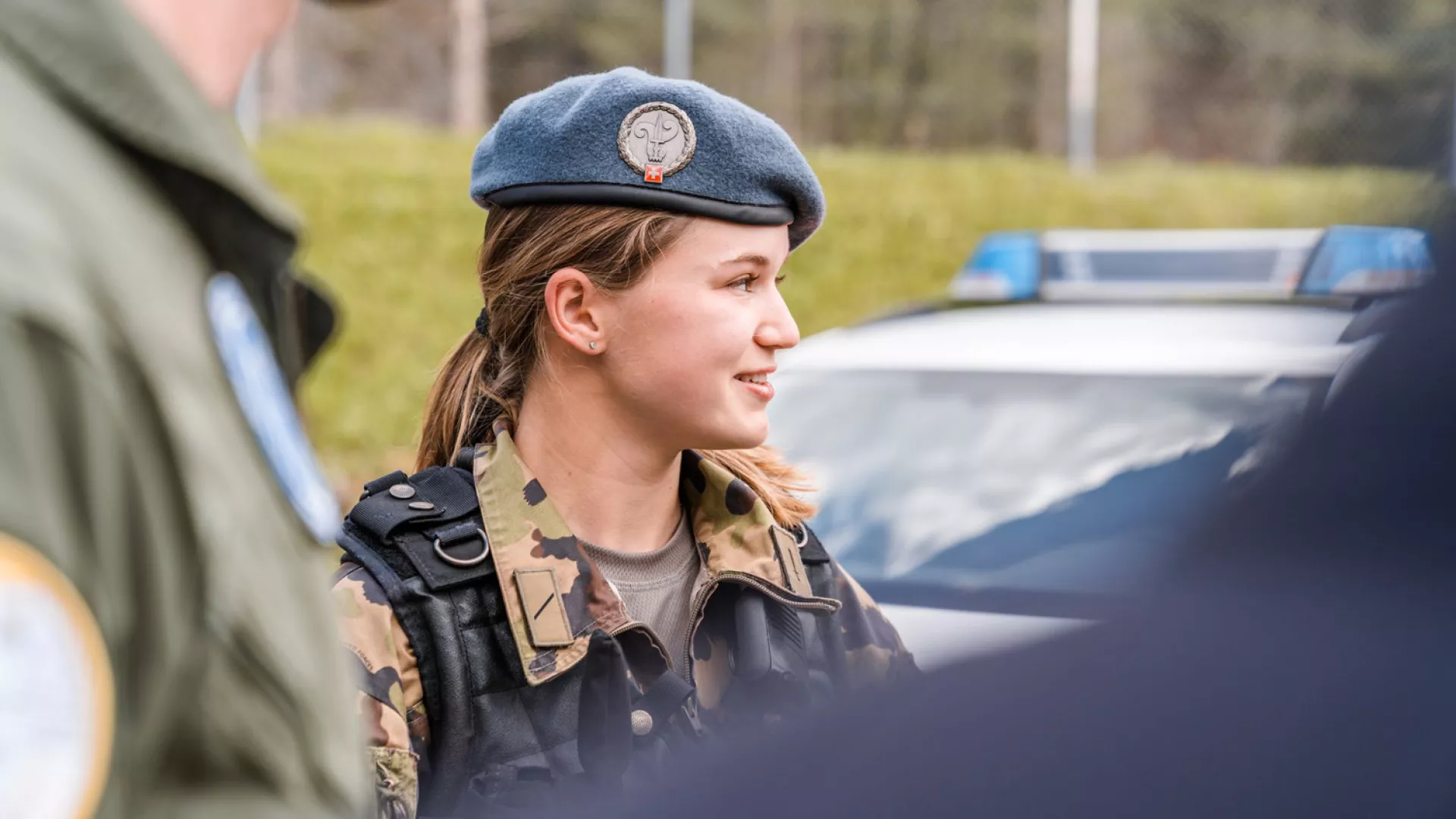 Jeune militaire en tenue de camouflage et béret échange lors d’une activité, avec un véhicule de service en arrière-plan