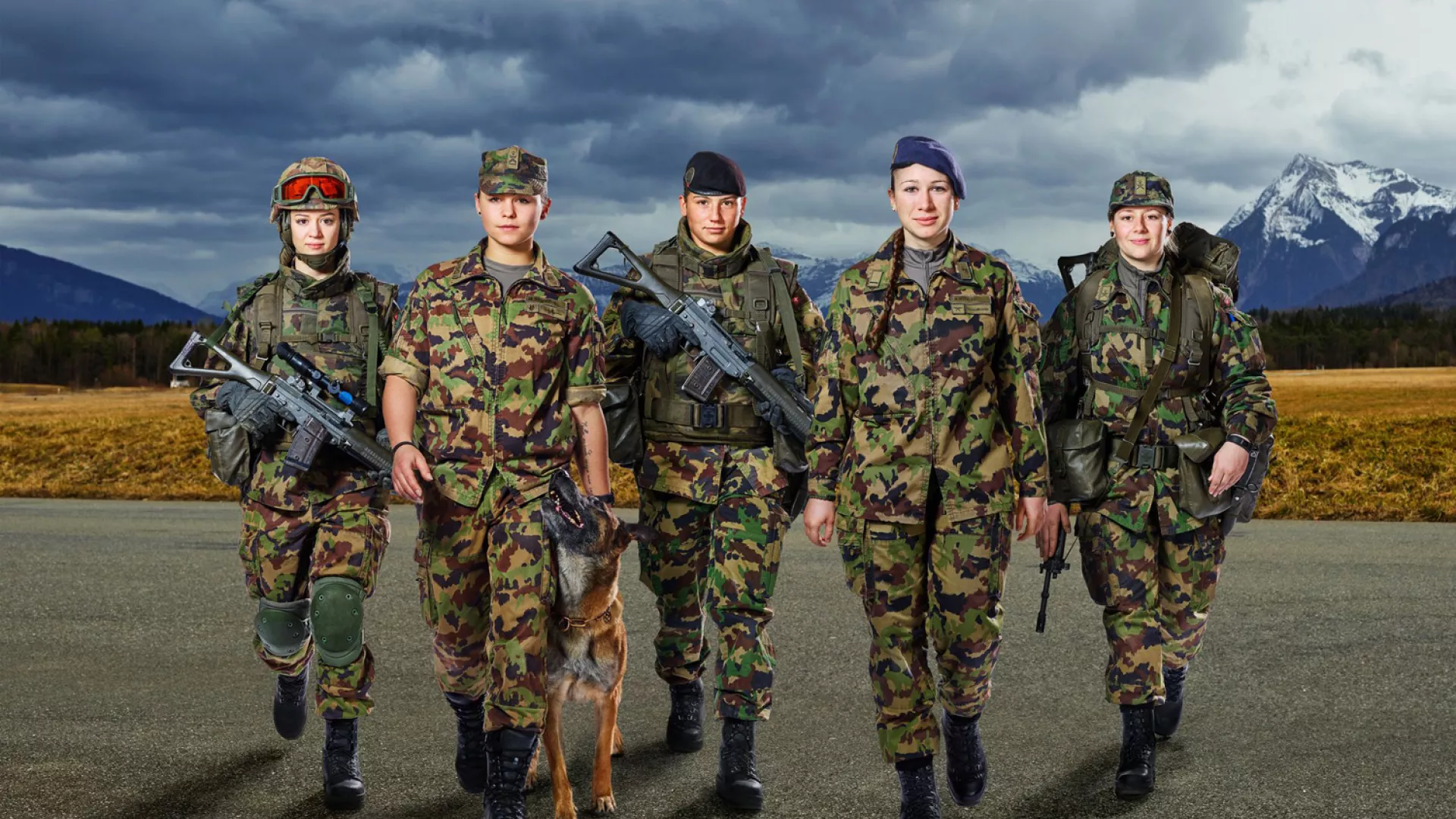 Groupe de femmes militaires en tenue de camouflage marchant côte à côte avec un chien, dans un paysage de montagne sous un ciel nuageux
