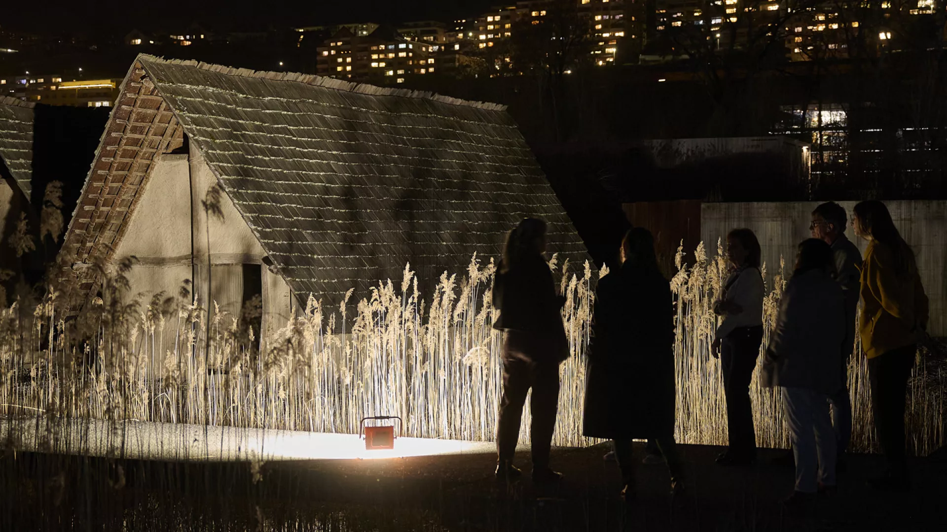 Personnes réunies de nuit près d’une reconstitution de maison préhistorique au Laténium.